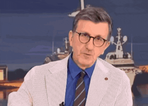 a man wearing glasses and a blue shirt is standing in front of a boat and says " einai ena kpatos komodia "