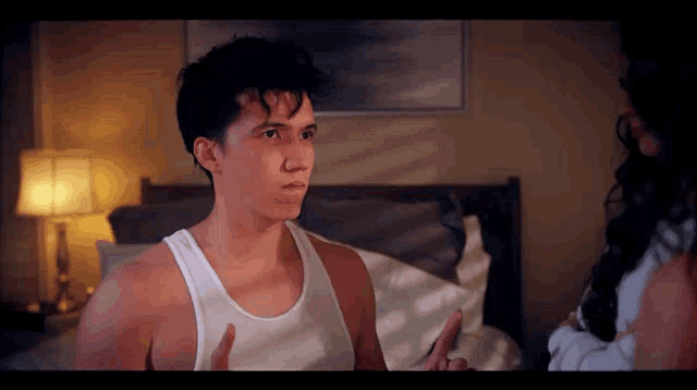 a man in a white tank top is talking to a woman in a bedroom
