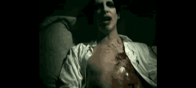 a man in a white shirt with blood on his chest is laying on a bed .