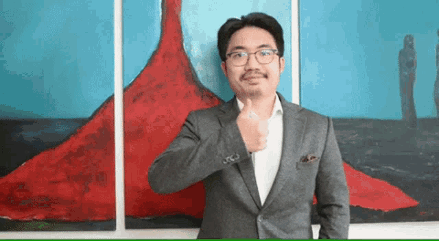 a man in a suit and glasses giving a thumbs up in front of a painting
