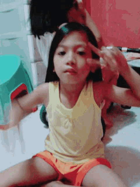 a little girl in a yellow tank top and orange shorts is sitting on the floor giving the peace sign .