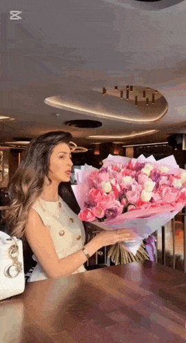 a woman is sitting at a table holding a large bouquet of pink flowers .