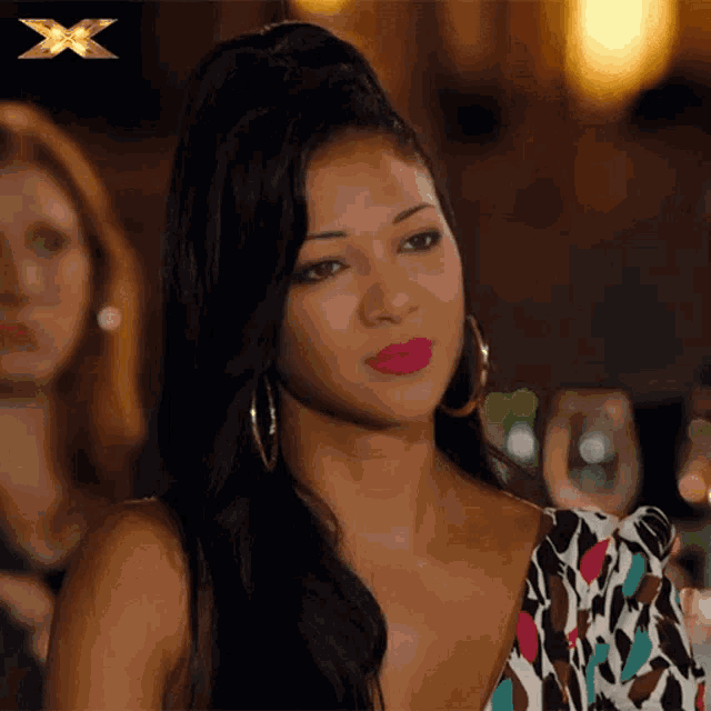 a woman with a ponytail and hoop earrings looks at the camera with an x in the background
