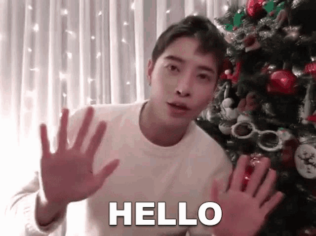 a young man is standing in front of a christmas tree and making a hello gesture with his hands .