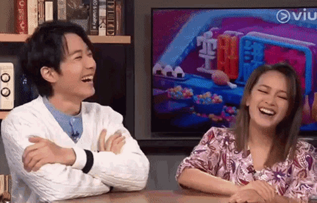 a man and a woman are sitting at a table and laughing in front of a television .