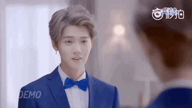 a young man wearing a blue suit and bow tie is looking at himself in the mirror .