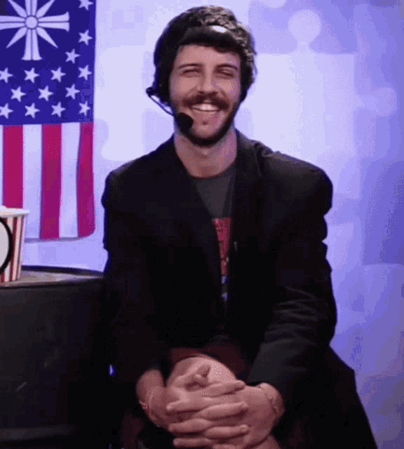 a man with a beard wearing a headset and smiling in front of an american flag