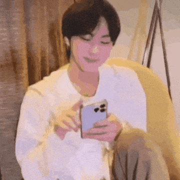 a man in a white shirt is sitting on a couch looking at his phone .