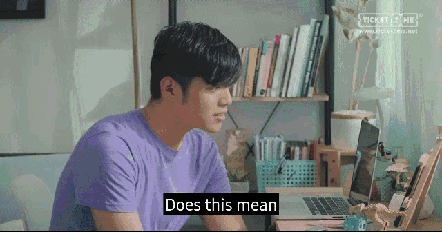 a man in a purple shirt is sitting at a desk in front of a laptop and says does this mean