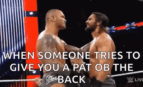 two wrestlers are hugging each other in a wrestling ring while someone tries to give you a pat on the back .