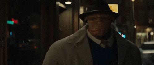 a man wearing a hat and a trench coat is walking down a street at night .