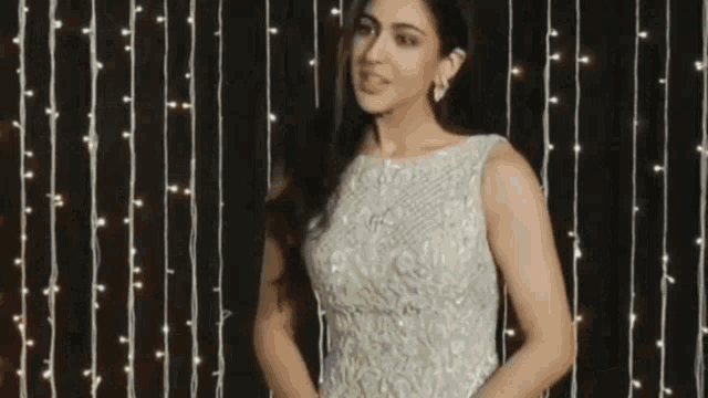 a woman in a white dress is standing in front of a string of lights and smiling .