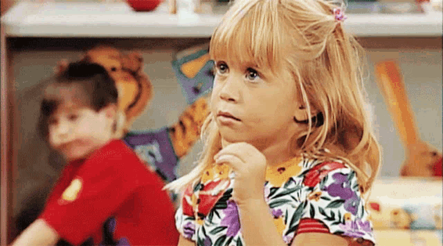 a little girl in a floral shirt looks at the camera with her hand on her chin