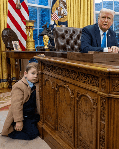 a little boy kneeling in front of a desk with donald trump sitting at it