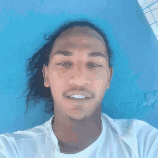 a man with long hair and a white shirt takes a selfie in front of a blue wall