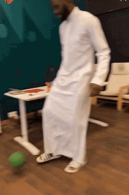 a man in a white robe is kicking a green ball on a wooden floor