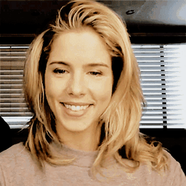 a woman with long blonde hair is smiling in front of a window with blinds