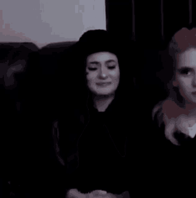 two women are sitting on a couch with their hands on their faces . one of the women is wearing a black hat .