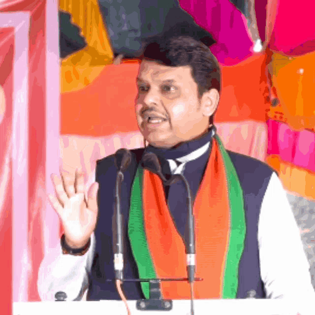 a man wearing an orange and green scarf is giving a speech in front of two microphones