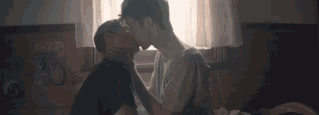 two young men are kissing in front of a window in a bedroom .
