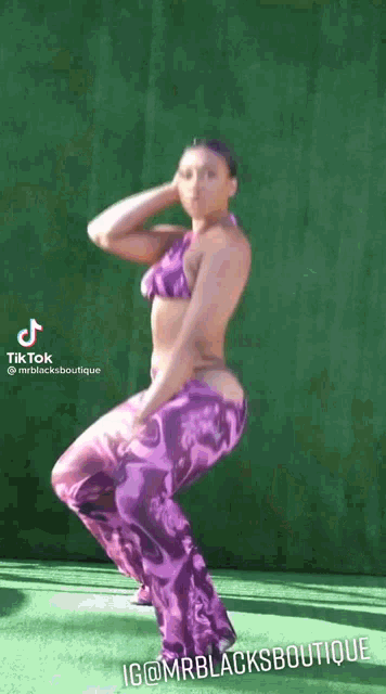 a woman in a purple bikini and purple pants is dancing in front of a green wall .