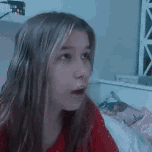 a young girl with long hair is sitting on a bed and making a surprised face .