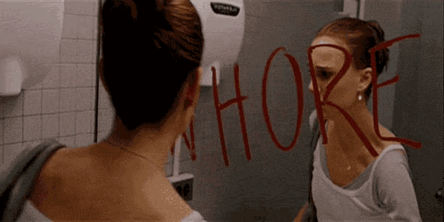 a woman is looking at herself in a bathroom mirror and has the word whore written on it .