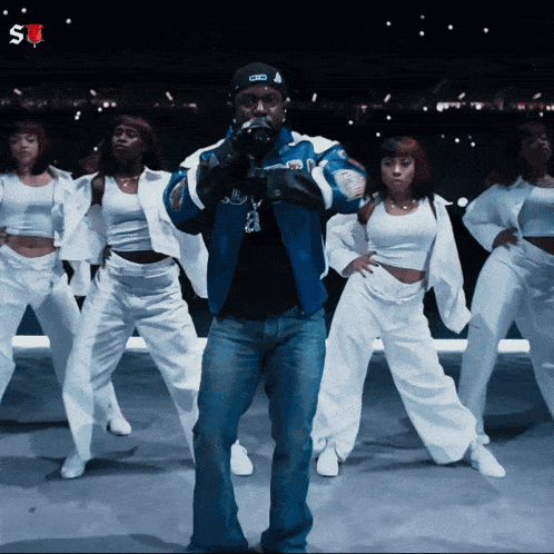 a man in a blue jacket is surrounded by female dancers
