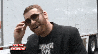 a man wearing sunglasses stands in front of a wwe.com exclusive trailer