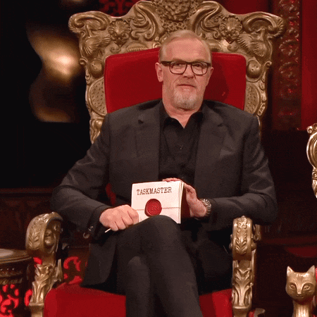 a man in a suit sits in a chair holding a book called taskmaster