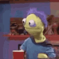 a green puppet with purple hair is holding a red cup and looking at the camera .