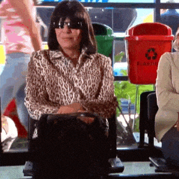 a woman wearing sunglasses and a leopard print shirt is sitting in a chair in front of a recycling bin .