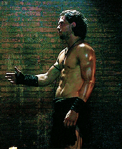 a shirtless man stands in front of a brick wall with his arm outstretched