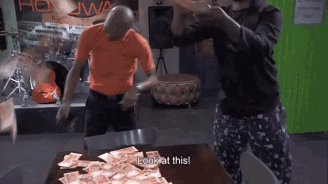 two men are standing in front of a table full of money and one of them is saying look at this