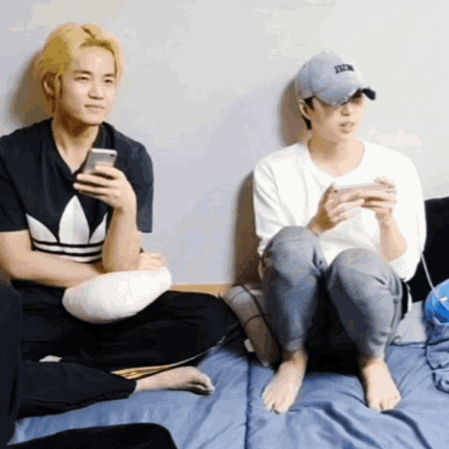 two young men are sitting on a bed looking at their phones .