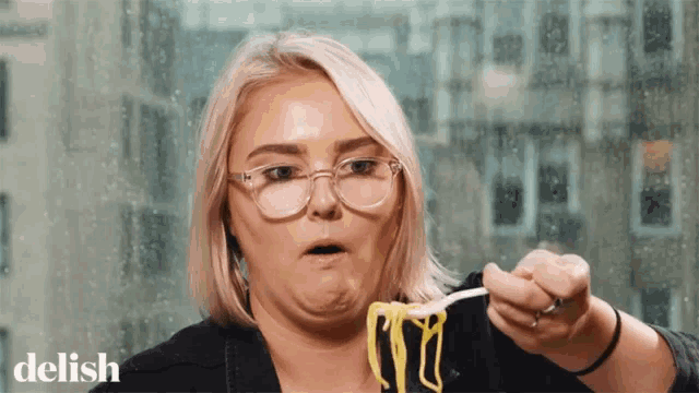 a woman wearing glasses is eating noodles with a fork and making a funny face .