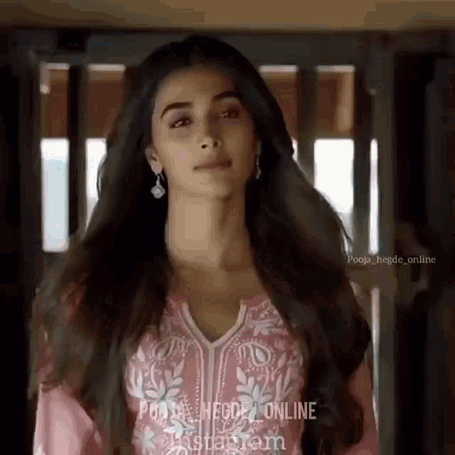 a woman with long hair is wearing a pink top and earrings and looking at the camera .