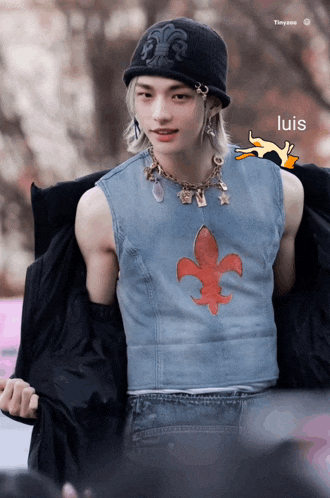 a young man wearing a beanie and a denim vest with a fleur de lis necklace
