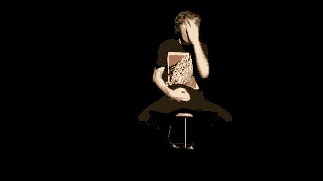 a man is sitting on a stool in a dark room with his hands on his hips .
