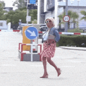 a woman wearing sunglasses is crossing a street in front of a blue sign with an arrow pointing left