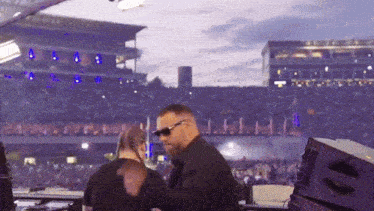 a man wearing sunglasses stands in front of a crowd at a stadium
