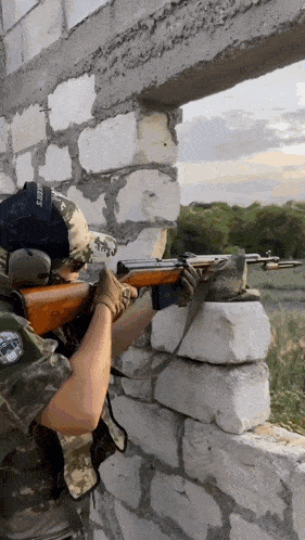 a man is holding a rifle in front of a brick wall with a hat that says ' sniper ' on it