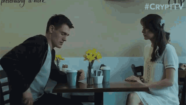 a man and a woman are sitting at a table with cups of coffee in front of a wall that says pretending to work