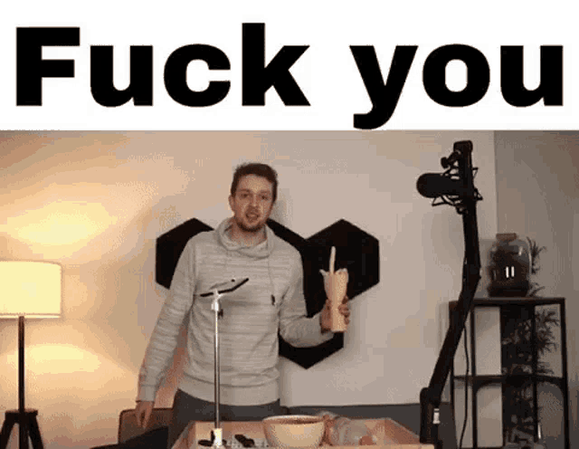 a man is standing in front of a microphone in a room holding a cup and giving the middle finger .