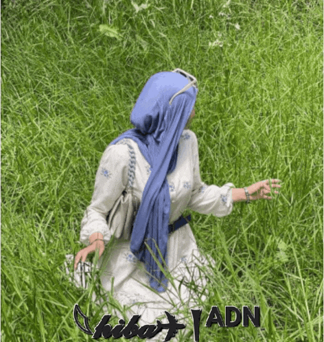 a woman in a blue scarf is kneeling in the grass with adn written on the bottom right