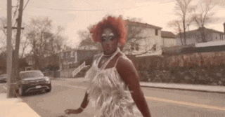 a drag queen is walking down a street wearing a white dress .