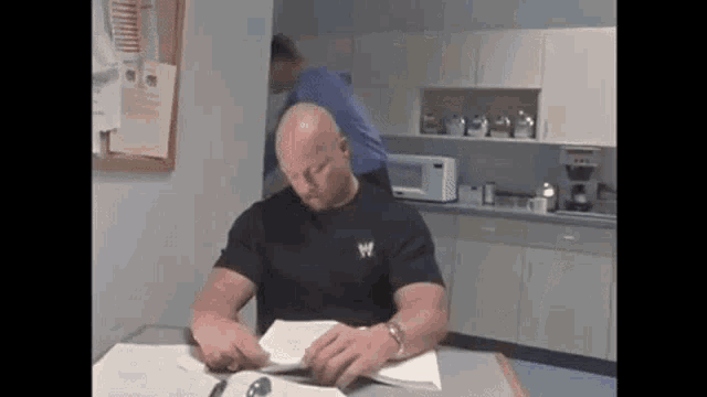 a man is sitting at a table reading a book while a man in a blue shirt stands behind him .