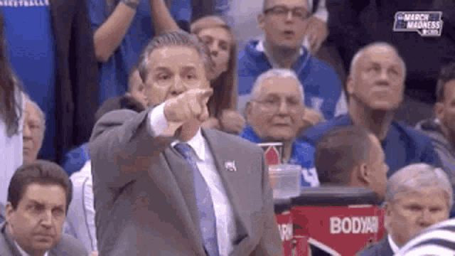 a man in a suit and tie is pointing at something while sitting in the stands of a basketball game .