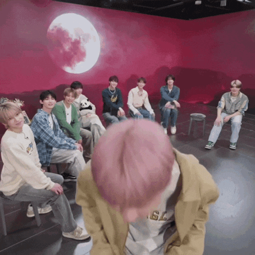a group of young men are sitting in a circle in front of a red wall with a full moon in the background