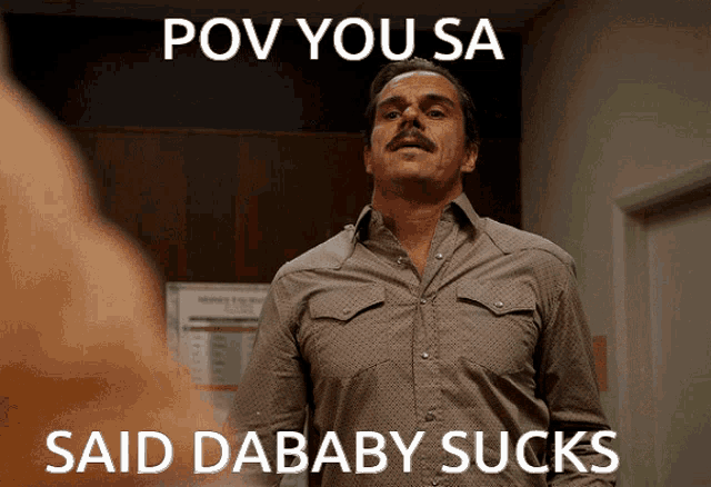 a man with a mustache stands in front of a sign that reads pov you sa said dababy sucks